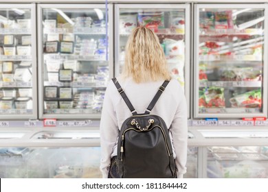 Young Woman At The Grocery Store In The Frozen Food Department. The Blonde In White Sweater With A Backpack. Back View.