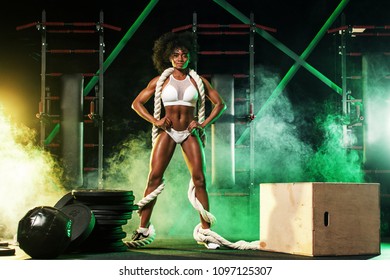 Young Woman In The Gym With A Battle Rope, In A Beautiful Sportswear, Against A Dark Background With Color Lights.