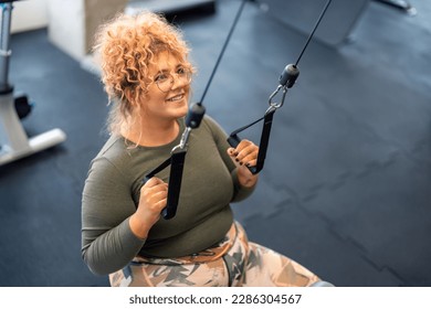 Young woman gym client with curly hair wearing sportswear motivated to exercise focused on her goal, doing cable pulldowns using cable pulley machine to workout her back and arms in gym fitness center - Powered by Shutterstock