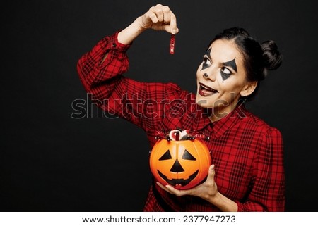 Young woman with Halloween makeup face art mask wear clown costume red dress holding Jack-o-Lantern carved pumpkin eating sweets isolated on plain black background studio. Scary holiday party concept