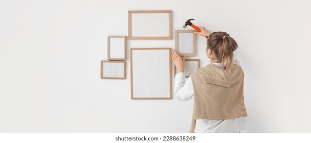 Young woman with hammer hanging blank photo frames on light wall