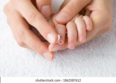 Young Woman Hand Holding Baby Hand And Applying Medical Ointment On Scratched Finger And Beaten Fingernail On White Towel. Mother Giving First Aid. Closeup. Front View. 