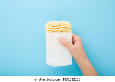 Young woman hand holding bright yellow dry noodles on light blue table background. Pastel color. Opened pack. Fast soup. Closeup. 