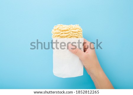 Young woman hand holding bright yellow dry noodles on light blue table background. Pastel color. Opened pack. Fast soup. Closeup. 