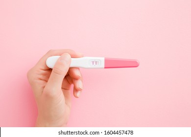 Young woman hand holding pregnancy test with two stripes. Pastel pink color background. Positive result. Closeup. Point of view shot. 