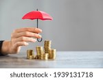 Young woman hand holding small red umbrella over pile of coins on table. Close up of stack of coins with female hands holding umbrella for protection. Financial safety and investment concept.  