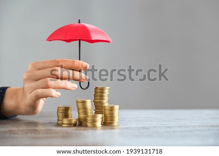 Young woman hand holding small red umbrella over pile of coins on table. Close up of stack of coins with female hands holding umbrella for protection. Financial safety and investment concept.  
