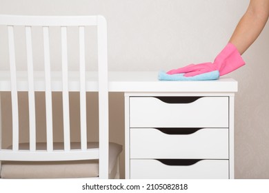 Young woman hand in pink rubber protective glove holding blue dry rag and wiping dust from white work table with drawers at home office. Closeup. General or regular cleanup in room. Cleaning service. 