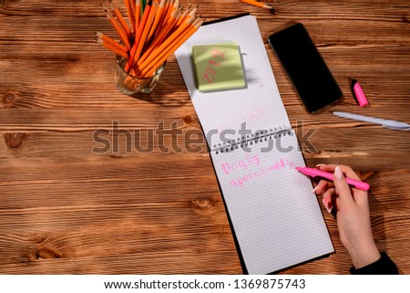 A young woman hands hold opened notebook pages with pink pencil in dark wooden table with bookmarks and telephone