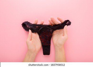 Young Woman Hands Holding Black Lace String Panties On Light Pink Table Background. Pastel Color. Daily Underwear. Closeup. Point Of View Shot.