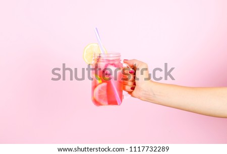Young woman hands holding freshly squeezed strawberry lemonade of citrus fruits. Female with mason jar full of cold cocktail, lemon, orange, lime & mint leaves. Pink background, copy space, close up.