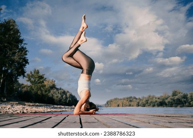 Young woman in handstand pose overlooking the river. Full length of a female yoga coach practicing yoga on the pier on riverside. Cloudy sky.