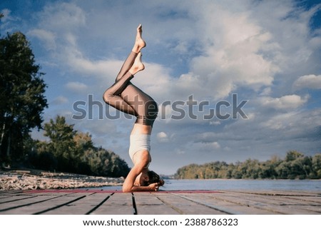 Young woman in handstand pose overlooking the river. Full length of a female yoga coach practicing yoga on the pier on riverside. Cloudy sky.