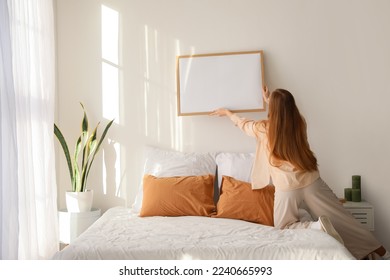 Young woman hanging blank frame on light wall in bedroom, back view - Shutterstock ID 2240665993