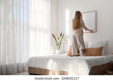 Young woman hanging blank frame on light wall in bedroom - Shutterstock ID 2248101117