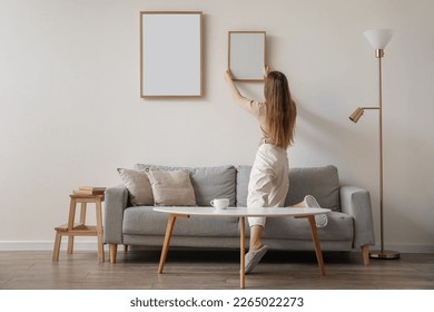 Young woman hanging blank frame on light wall at home, back view - Shutterstock ID 2265022273