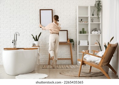 Young woman hanging blank frame on white brick wall in bathroom, back view