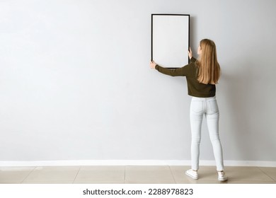 Young woman hanging blank frame on light wall at home, back view