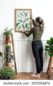 Young Woman Hanging A Frame Of Leaf Print Painting On The Wall