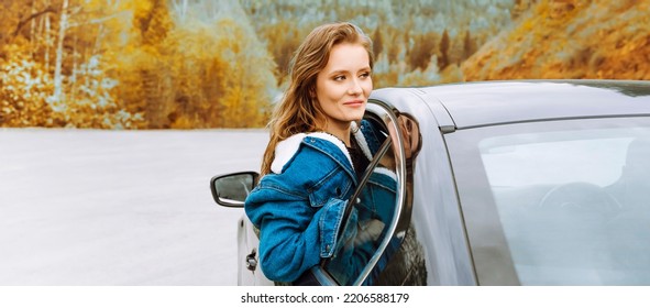 Young Woman Hanging Out Of A Car Window And Looking At The Road Ahead, With Autumnal Trees In The Background