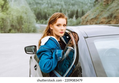 Young Woman Hanging Out Of A Car Window And Looking At The Road Ahead, With Autumnal Trees In The Background