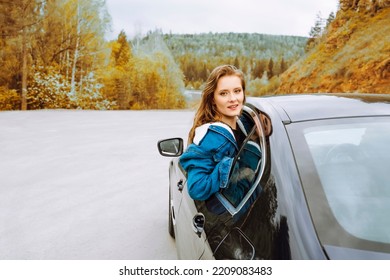 Young Woman Hanging Out Of A Car Window And Looking At The Road Ahead, With Autumnal Trees In The Background