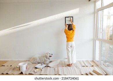 Young woman hanging picture frame in room, decorating her newly renovated apartment, stands with her dog in white room