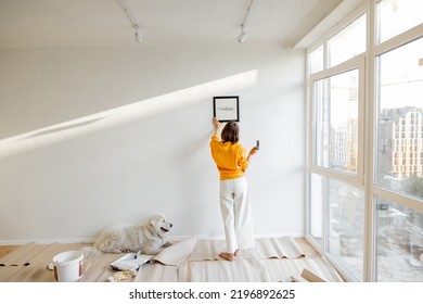 Young woman hanging picture frame in room, decorating her newly renovated apartment, stands with her dog in white room - Shutterstock ID 2196892625