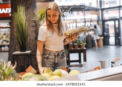 Young woman happily choosing a fresh pineapple at a grocery store. Surrounded by vibrant colors and textures. Her enthusiasm for healthy eating shines through as she carefully considers her options - Powered by Shutterstock