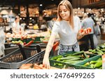 Young woman happily shops for fresh produce in a grocery store, selecting tomatoes and cucumbers for a healthy lifestyle. The vibrant vegetables stand out against a blurred background of shoppers