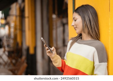 Young woman happily using smartphone against yellow city wall, embodying tech savvy generation z lifestyle outdoors - Powered by Shutterstock