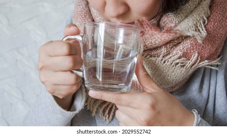 A young woman has a cold and drinks medicine. The girl her throat with a scarf and drank hot water. Treatment with folk remedies. A lot of copious drinking is useful during a cold. - Shutterstock ID 2029552742