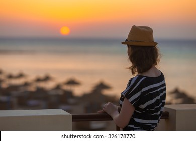 Young woman in hat and cute summer dress standing at the terrace with peaceful sea scenery, looking at sunset or sunrise on horizon, back view, copy space
