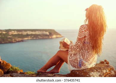 Young woman in hat and cute summer dress standing on the stone, looking at sunset over the sea