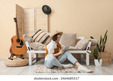 Young woman in hat and eyeglasses using tablet and sitting on floor in living room - Powered by Shutterstock