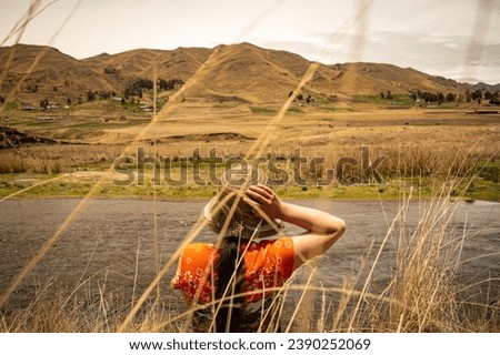 young woman with hat in the foreground enjoying the landscape, nearby river, distant mountains and cloudy sky in the background in Langui, Cusco, Peru.