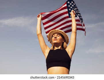 A Young Woman In A Hat Holds The US Flag Against The Blue Sky. Portrait Of A Patriot Woman. American Holiday