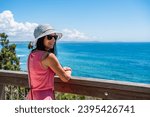 A young woman in a hat and a pink dress stands smiling at the railing on the observation deck and looks at the seaside landscape on a sunny summer day.