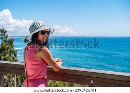 A young woman in a hat and a pink dress stands smiling at the railing on the observation deck and looks at the seaside landscape on a sunny summer day.