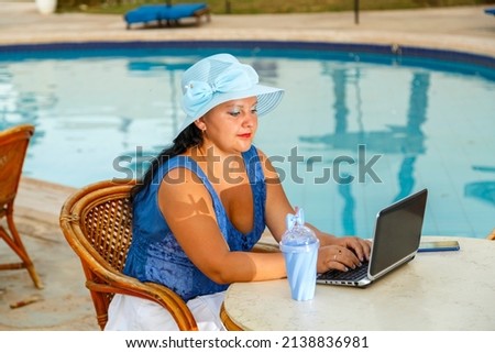 A young woman in a hat at a table in the morning next to the pool and works remotely on a laptop. Horizontal photo