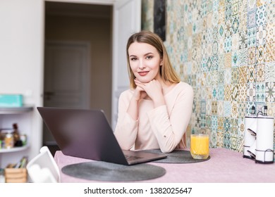Young woman having breakfast in the kitchen, working laptop and drinking an healthy orange juice