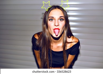 Young woman having fun, showing long tongue, going crazy alone, bright sexy make up, funny party hairs accessory with stars, night image with flash.
