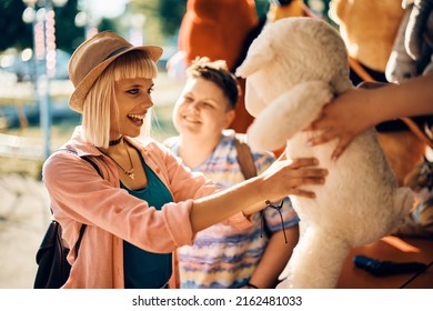 Young Woman Having Fun While Getting Plush Toy At Carnival Both After Winning In Target Shooting Game With Her Girlfriend At Amusement Park. 