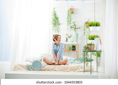 Young Woman Having A Good Morning Sitting On The Bed In The Beautiful Bright Bedroom With Green Plants And Clock