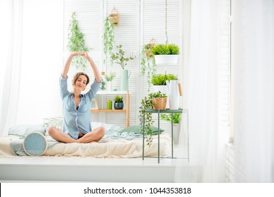 Young Woman Having A Good Morning Sitting On The Bed In The Beautiful Bright Bedroom With Green Plants And Clock