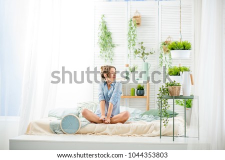 Young woman having a good morning sitting on the bed in the beautiful bright bedroom with green plants and clock