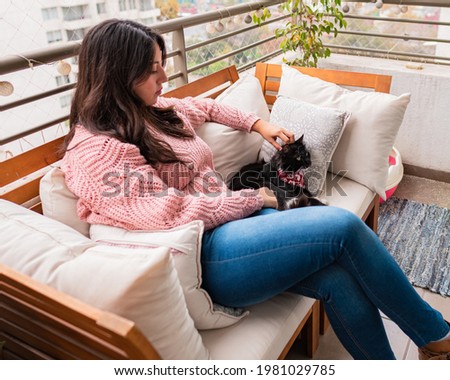 Young woman having a good time at her couch in the balcony with her black cat dressing a red cravat