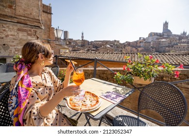 Young woman having lunch with pizza and wine at outdoor restaurant with beautiful view on the old town of Siena. Concept of italian cuisine and traveling Tuscany region of Italy