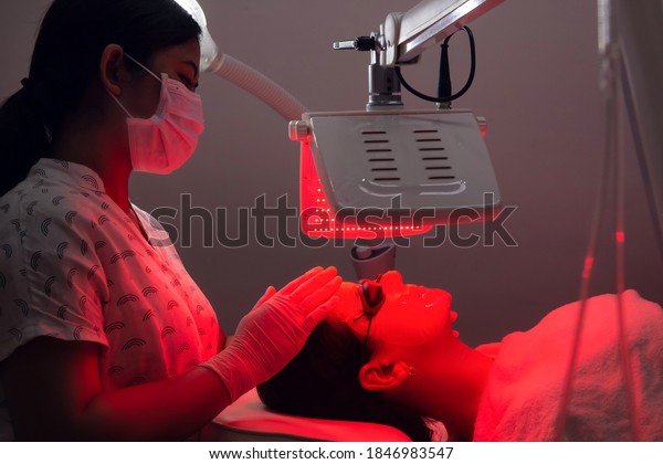 Young woman having red LED light facial therapy\
treatment in beauty salon. Beautician wearing face mask maintaining\
safety procedures during appointment. Beauty, new normal and\
wellness concept