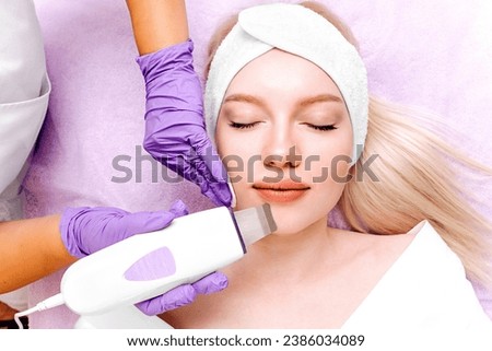 Young woman with a headband receives an ultrasonic peeling treatment on her face. Cosmetologist exfoliates the skin with an ultrasonic spatula in a beauty salon. Ultrasonic skin cleansing procedure. 